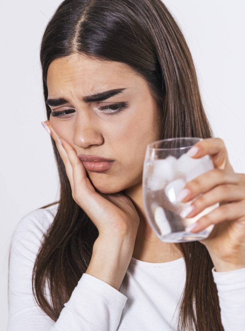 Young woman with sensitive teeth and hand holding glass of cold water with ice. Healthcare concept. woman drinking cold drink, glass full of ice cubes and feels toothache, pain